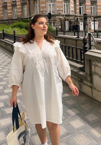 Λευκό βαμβακερό φόρεμα με φουσκωτά μανίκια, βολάν και κεντημένα διακοσμητικά στο λαιμό. Άτομο κρατά μια τσάντα tote με σκούρους μπλε λαβές.