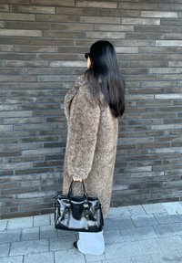 Καφέ, φουντωτός μανδύας με πλήρη μανίκια; μαύρη δερμάτινη τσάντα με ασημένιες λεπτομέρειες; γκρι παντελόνι; στέκεται μπροστά σε δομημένο τοίχο από πέτρα.