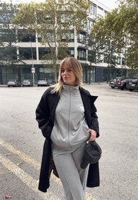 Harmaa urheilupuku, jossa on vetoketjulla varustettu takki ja siihen sopivat housut, joissa on valkoiset raidat sivuilla. Asusteena pitkä musta takki ja musta lippalakki.