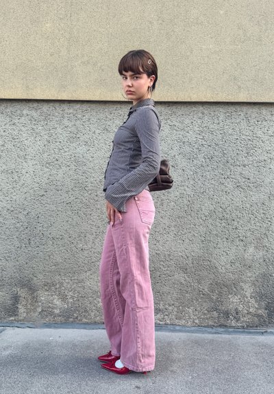 Camisa de rayas de manga larga, pantalones rosas de pierna ancha, zapatos rojos de punta. Bolsa marrón, pendientes de aro, fondo de pared gris texturizado.