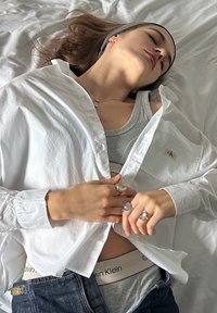 Hvit skjorte med knapper delvis åpnet over en grå singlet, med denimjeans. Flere sølvringer pryder hendene. Mykt sengetøy i bakgrunnen.