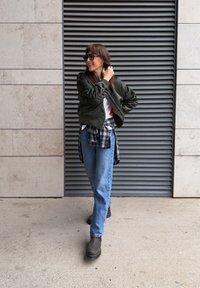 Blouson aviateur vert olive, t-shirt blanc, chemise à carreaux nouée à la taille, jean bleu, bottines marron montantes ; debout devant un mur gris ventilé.
