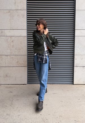 Chaqueta bomber verde oliva, camiseta blanca, camisa de cuadros atada a la cintura, vaqueros azules, botines marrones; de pie frente a una pared gris con rejillas.