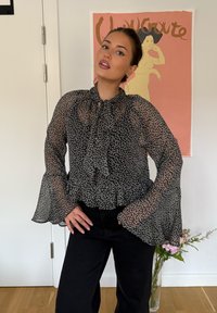Schwarze, transparente Bluse mit kleinem weißen Blumenmuster, gerafftem Saum und langen Glockenärmeln, kombiniert mit dunklen Jeans. Blumenmuster im Hintergrund.