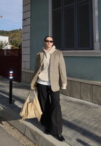 Oseba z sončnimi očali, bež kapuco, karo suknjičem, črnimi hlačami in črnimi čevlji, ki drži bež torbo z resicami na sončni pločniku.