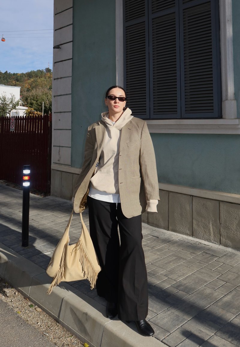 Isik kandmas päikeseprille, beeži kapuutsiga pusa, ruudulist jopet, musti pükse ja musti kingi, hoides päikeselisel kõnniteel beežist tuttidega kotti.