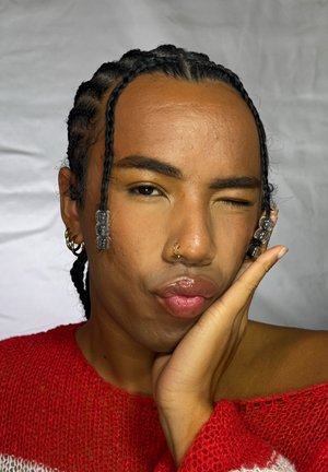 Persona con cabello trenzado, cuentas transparentes, pendientes en la nariz y las orejas, vistiendo un suéter rojo de punto, guiñando un ojo con los labios fruncidos y la mano apoyada en la mejilla.