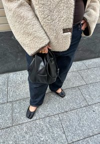 Isik, kes kannab beežikat pehmet jopet, tumeid teksapükse, musti jagatud varbaga kingi ning hoiab musta nahast ämbrikujulist kotti, seistes hallil plaatidega sillutatud tänaval.