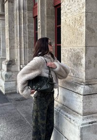 Beige faux fur jacket, black leather handbag with silver fittings, camouflage trousers, and dark sunglasses; leaning against a stone wall.