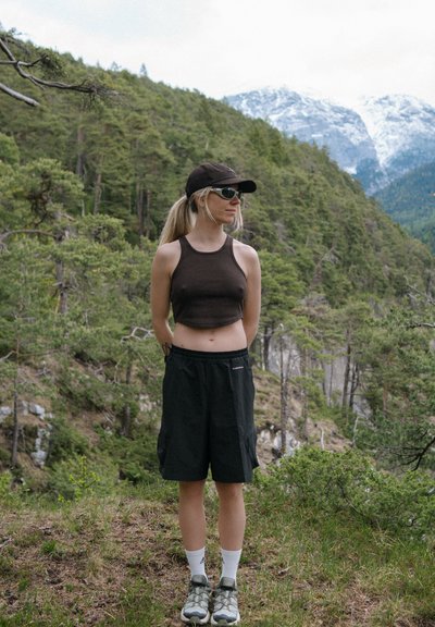 Joven mujer con gorra negra, gafas de sol, top corto y pantalones cortos, de pie en un bosque con montañas nevadas al fondo.