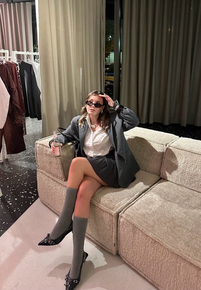 Blazer gris de gran tamaño sobre una camisa blanca, falda negra y calcetas grises hasta la rodilla. Sentada en un sofá de color crema texturizado, frente a suaves cortinas.