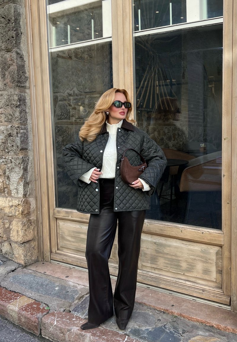 Femme portant une veste matelassée, un pull blanc, un pantalon en cuir noir, des lunettes de soleil, et tenant un sac à main marron, debout près d'un mur en pierre et d'une porte en verre.