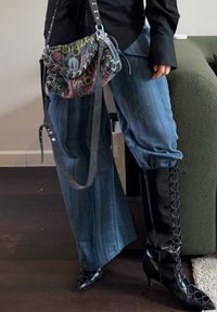 Personne portant un jean bleu ample rentré dans des talons hauts noirs à lacets, un haut noir à manches longues, portant un sac à bandoulière coloré avec du texte.