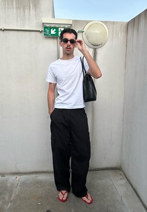 Hombre ajustándose las gafas de sol, vistiendo una camiseta blanca, pantalones cargo negros, chanclas rojas y llevando una bolsa de mano negra contra paredes de hormigón.