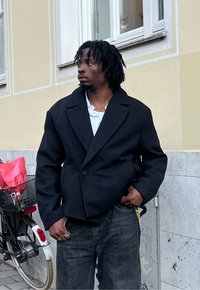 Czarny, oversize'owy płaszcz z szerokim kołnierzem i zapinany na pojedynczy guzik, noszony na białej koszuli, zestawiony z ciemnymi dżinsami.