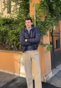 Giubbotto imbottito di colore blu scuro con il marchio "POLO CLUB", indossato sopra una camicia azzurra chiara. Pantaloni beige e scarpe scure, in piedi davanti a un ambiente verde.