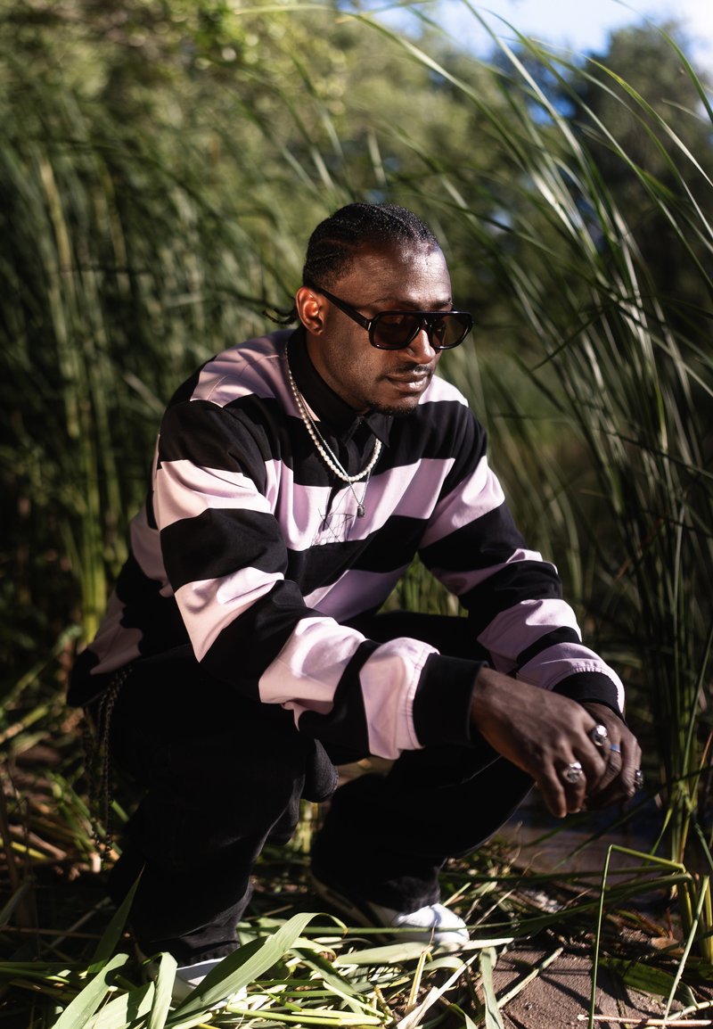 Homme portant des lunettes de soleil, une chemise rayée noire et rose, et des bijoux, accroupi en plein air parmi une herbe verte et haute.