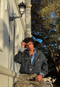 Chaqueta de mezclilla con un corte relajado, camisa de rayas azul y blanca debajo, pantalones de camuflaje y auriculares negros. Entorno al aire libre con árboles.