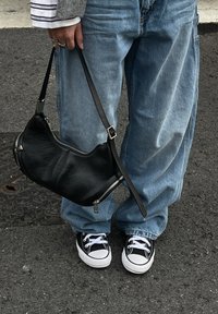 Mala de ombro em couro preto com fechos de correr, segurada por uma pessoa vestindo jeans azuis soltos e ténis pretos com detalhes brancos. Superfície texturizada visível.