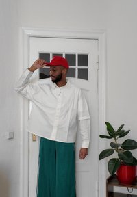 Cămașă albă supradimensionată, cu un tiv rotunjit, asortată cu pantaloni largi de culoare teal. O șapcă roșie se află pe cap, alături de o plantă în ghiveci.