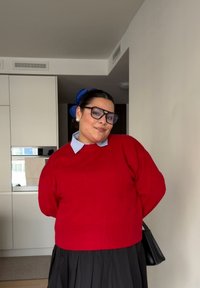 Crveni pleteni džemper s rebrastim rubom preko svijetloplave košulje s ovratnikom, u kombinaciji s crnom plisiranom suknjom i crnom torbicom.