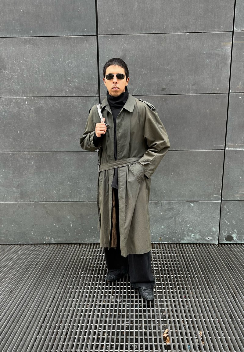 Homem vestindo óculos de sol e um longo casaco de trench coat olival, em pé sobre uma grade metálica contra uma parede de azulejos cinzentos.