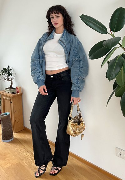 Chaqueta bomber azul clara, camisa blanca ajustada, pantalones vaqueros negros de campana, sandalias negras, bolso dorado. Entorno interior con suelo de madera y plantas.