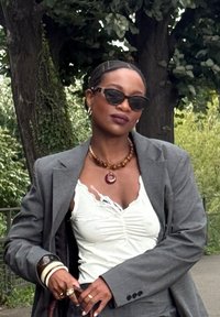 Grau maßgeschneiderter Blazer mit leichtem Innenfutter, weißem Rüschenshirt, bernsteinfarbener Halskette und schwarzen Sonnenbrillen, vor einer grünen Kulisse.