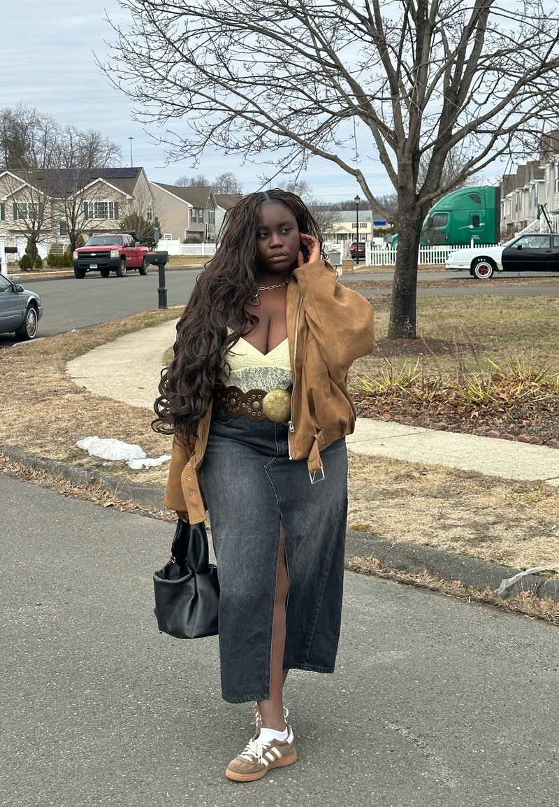 Jeune femme aux longs cheveux bouclés marchant dans une rue de banlieue, portant une veste beige, un haut jaune, une jupe foncée fendue, des baskets, et portant un sac noir.