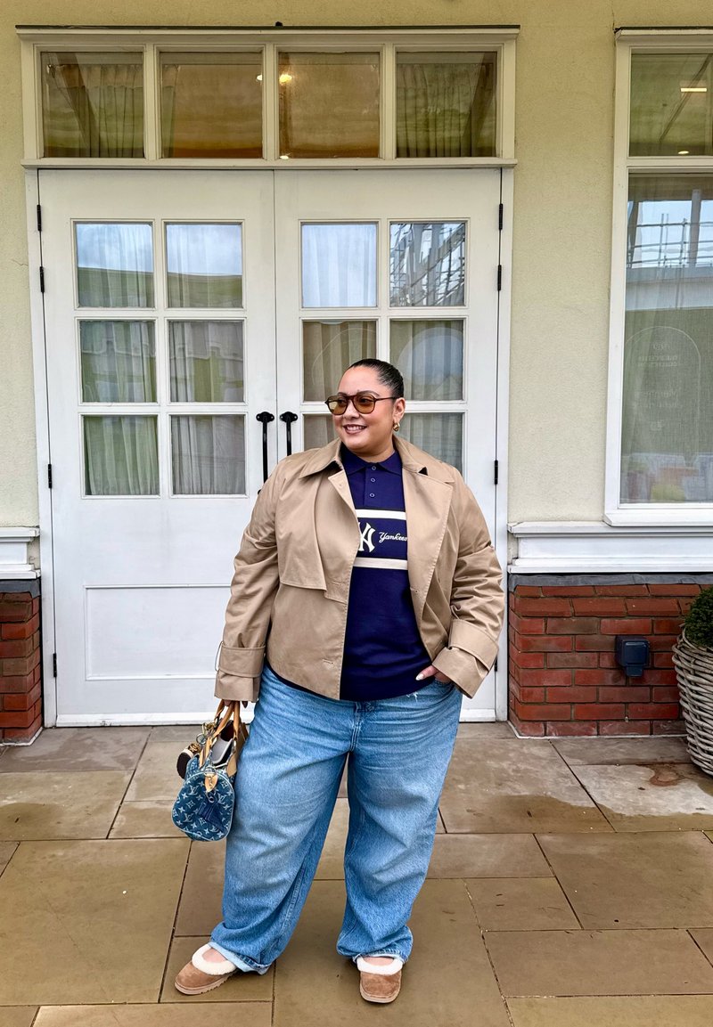 Personne portant une veste beige, une chemise bleu marine, un jean bleu et des lunettes de soleil, se tient devant une porte blanche en tenant un sac à main bleu.