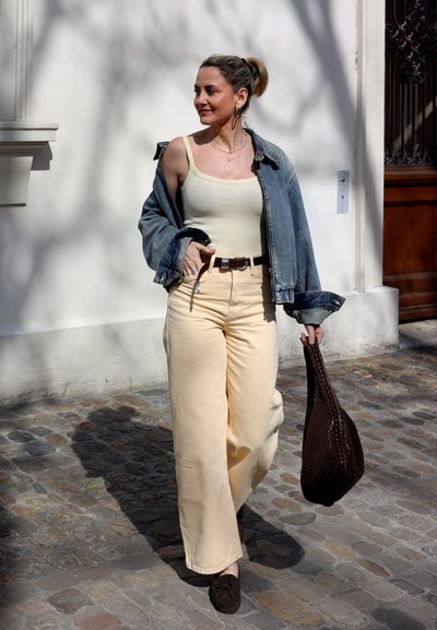 Mujer caminando por una calle empedrada con pantalones beige, camiseta sin mangas crema, chaqueta vaquera caída de los hombros, sosteniendo un bolso marrón tejido.