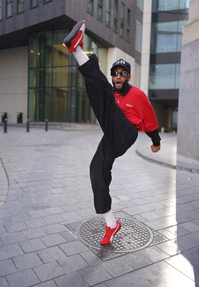 Camisa roja de gran tamaño con mangas negras, pantalones anchos negros, zapatillas rojas y calcetines blancos. Pierna levantada, de pie sobre una tapa de alcantarilla.