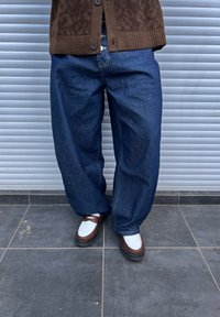 Pantaloni din denim de culoare bleu închis, cu picioare largi și cusături vizibile, asortați cu loafers maro și albi pe un podea texturată de culoare neagră, pe un fundal gri.