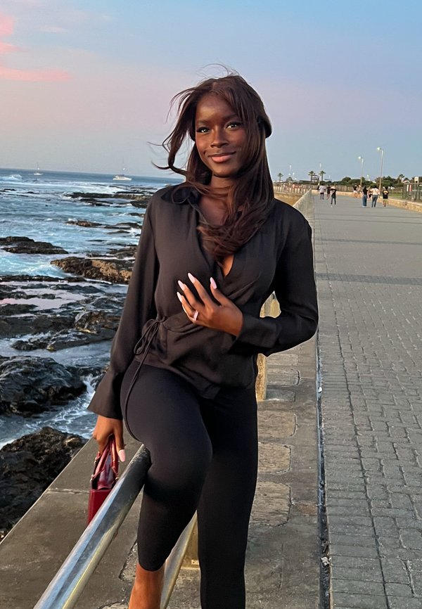 Svart långärmad blus med knytning och djup urringning, kombinerad med svarta leggings. En röd handväska ger en färgklick till outfiten. Havet i bakgrunden.