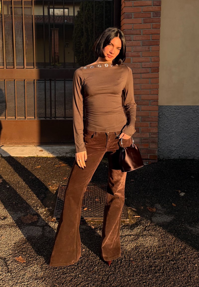 Haut marron à manches longues avec des boutons décoratifs à l'épaule, associé à un pantalon évasé marron à texture mate, tenant un petit sac à main noir.