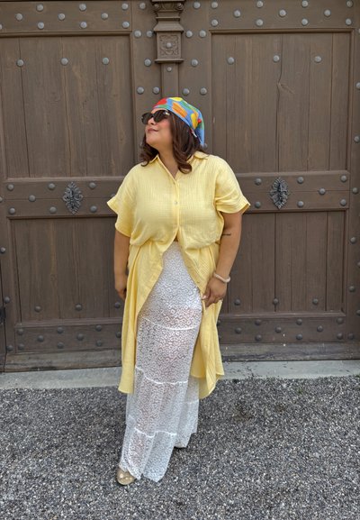 Mujer con un pañuelo de cabeza colorido, gafas de sol, camisa amarilla abotonada sobre una falda de encaje blanca, de pie frente a una gran puerta de madera con clavos de metal.