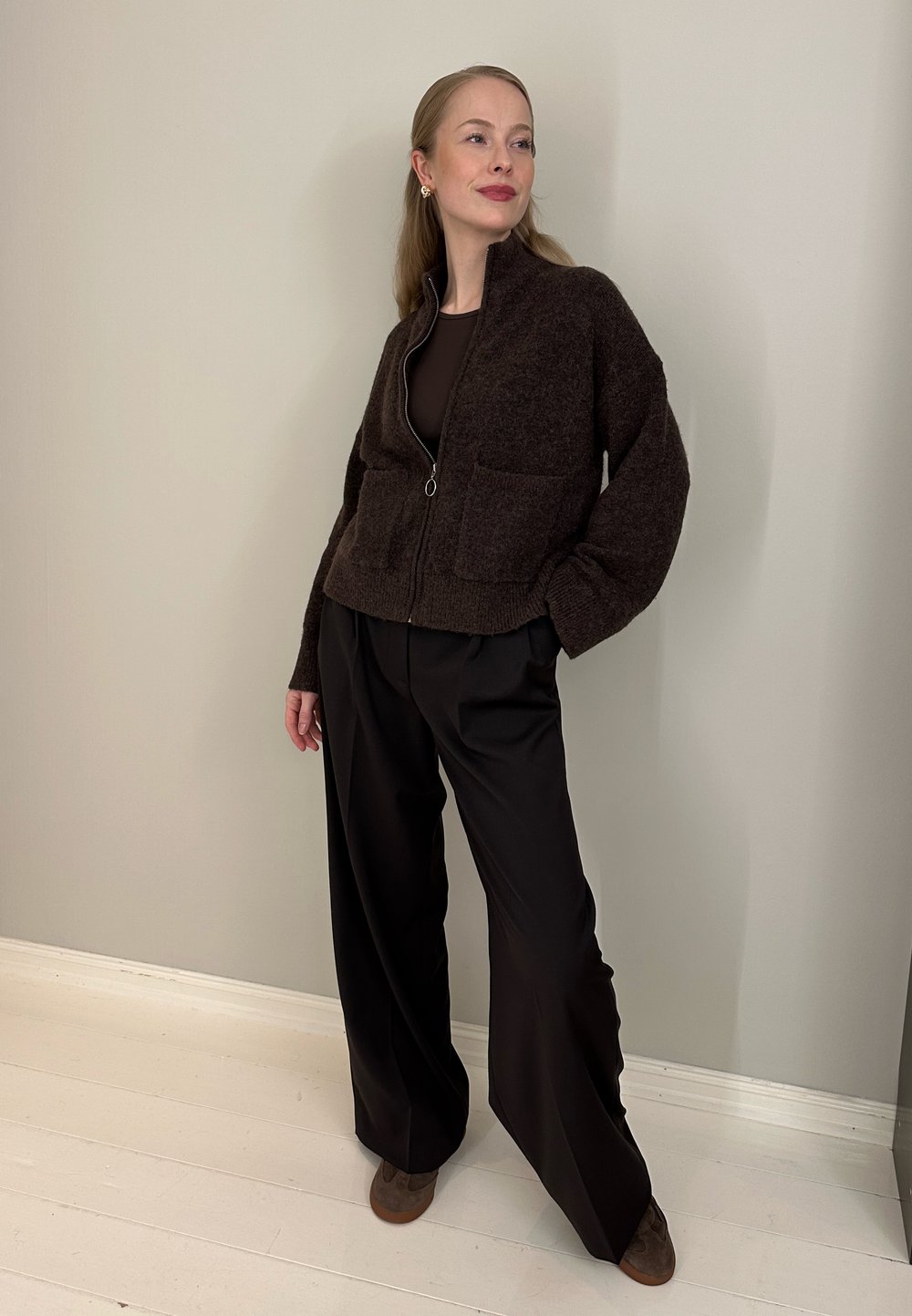 Femme debout à l'intérieur portant un cardigan zippé marron foncé, un pantalon large noir et des chaussures marron, devant un mur beige uni.
