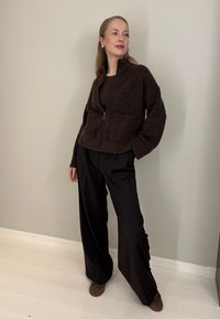 Femme debout à l'intérieur portant un cardigan zippé marron foncé, un pantalon large noir et des chaussures marron, devant un mur beige uni.