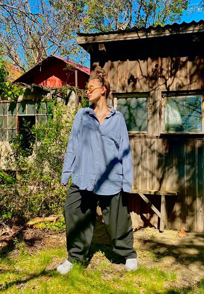 Camisa azul claro de gran tamaño con cuello abotonado, combinada con pantalones anchos negros y zapatos grises sin cordones, situada en un fondo natural.