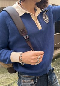 Niebieski sweter dziany z kremowym kołnierzem, z naszywką z logo. Ręka ozdobiona srebrnymi pierścieniami i teksturowaną bransoletką. Widoczny brązowy pasek.