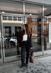 Μαύρα παντελόνια φαρδιά, άσπρο φανελάκι, υπερμεγέθη μαραντζίν jacket και μαύρα παπούτσια; κρατώντας μια καφέ τσάντα ώμου με κείμενο. Ο γυάλινος τοίχος αντανακλά τα γύρω.