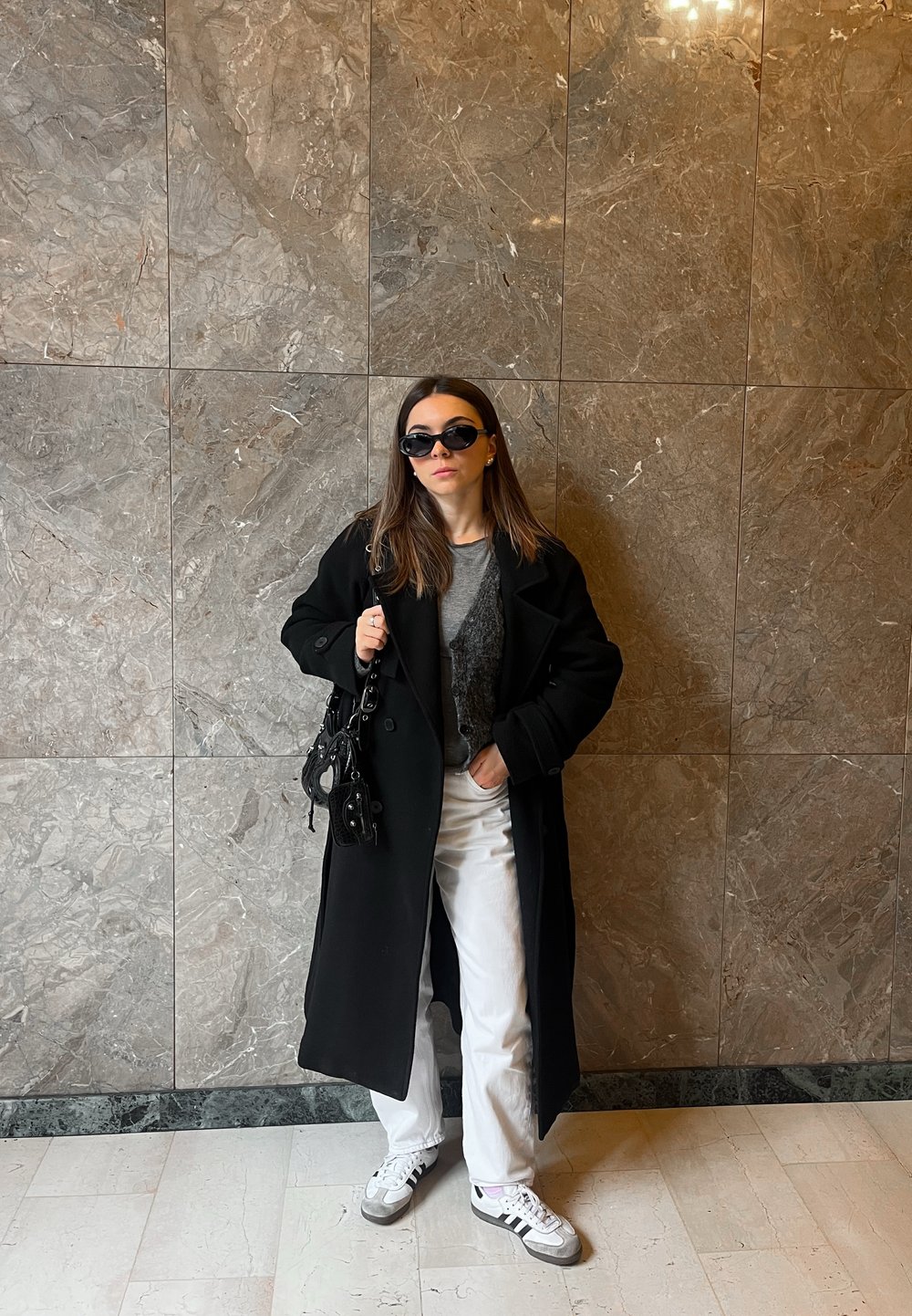 Zwarte wollen jas, grijze gebreide top, witte jeans, zwarte handtas en sneakers met grijze accenten. Zonnebril, marmeren muur als achtergrond, binnenomgeving.