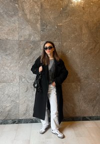 Zwarte wollen jas, grijze gebreide top, witte jeans, zwarte handtas en sneakers met grijze accenten. Zonnebril, marmeren muur als achtergrond, binnenomgeving.