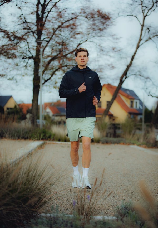 Chaqueta con capucha negra con el logo de Nike, pantalones cortos verdes claros, calcetines blancos y zapatillas de correr, con un fondo suburbano.
