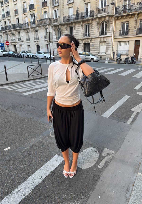 Femme aux cheveux foncés, portant un haut blanc et un pantalon noir, ajuste ses lunettes de soleil en se tenant dans une rue de Paris près d'un passage piéton avec des voitures garées.