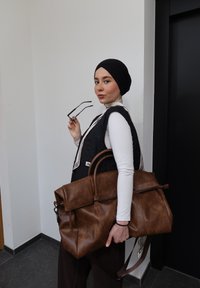 Jeune femme portant un foulard noir et un gilet, tenant des lunettes et un grand sac en cuir marron, debout à l'intérieur contre des murs blancs et noirs.