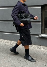 Borsa a tracolla in pelle nera con hardware argentato, camicia oversize viola scuro, pantaloni shorts neri al ginocchio e scarpe nere con calzini.