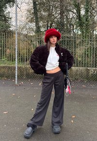 Jeune femme debout en plein air, portant une veste en velours côtelé noire, une chemise blanche, un pantalon noir à jambes larges, des sabots noirs et un chapeau rouge duveteux, tenant un sac avec des porte-clés.