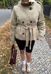 Cappotto trench beige doppiopetto con cintura, dettagli di bottoni e spalline, abbinato a pantaloni shorts neri per bicicletta e sneakers beige.