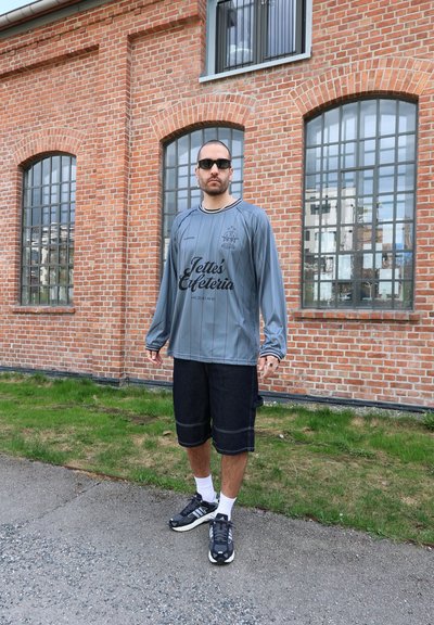 Jersey gris de manga larga con rayas negras y logo, combinado con pantalones cortos de mezclilla oscura y zapatillas deportivas negras, contra un muro de ladrillo.